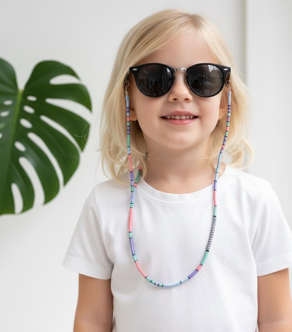 A child wearing black round sunglasses with a colorful pastel wrapped rope strap strap is fully visible, smiling, with a white t-shirt and a monstera plant in the background.